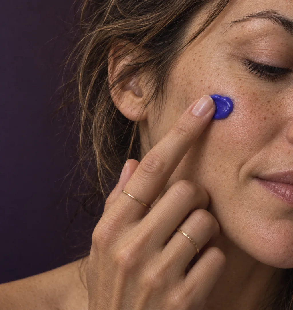 Illustrative close-up of a freckled woman with natural brows and fine gold rings applying a small dab of violet cream.